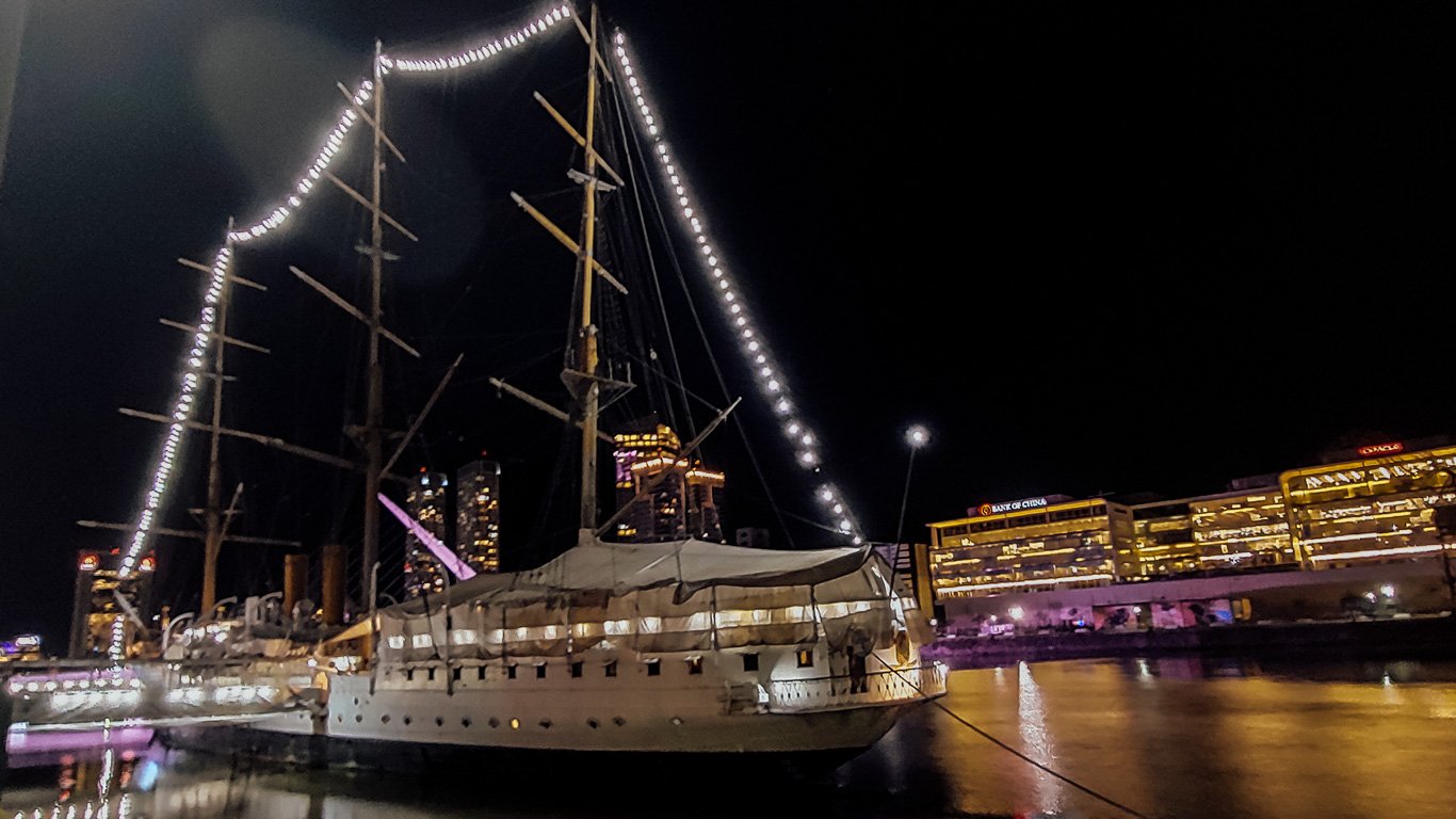 Fragata-Sarmiento Navio Fragata Sarmiento ancorado em Puerto Madero, iluminado por luzes ao longo dos mastros, refletindo nas águas calmas. Prédios comerciais brilham ao fundo no contraste da noite.