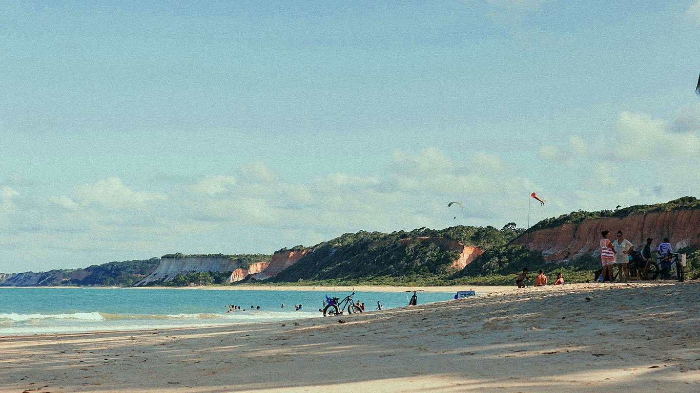 Praia-da-Pitinga Praia da Pitinga, com areia dourada, falésias avermelhadas ao fundo e mar esverdeado compõem a paisagem paradisíaca da Praia da Pitinga. Pessoas relaxam, algumas se aventuram no parapente e outras se refrescam na água.