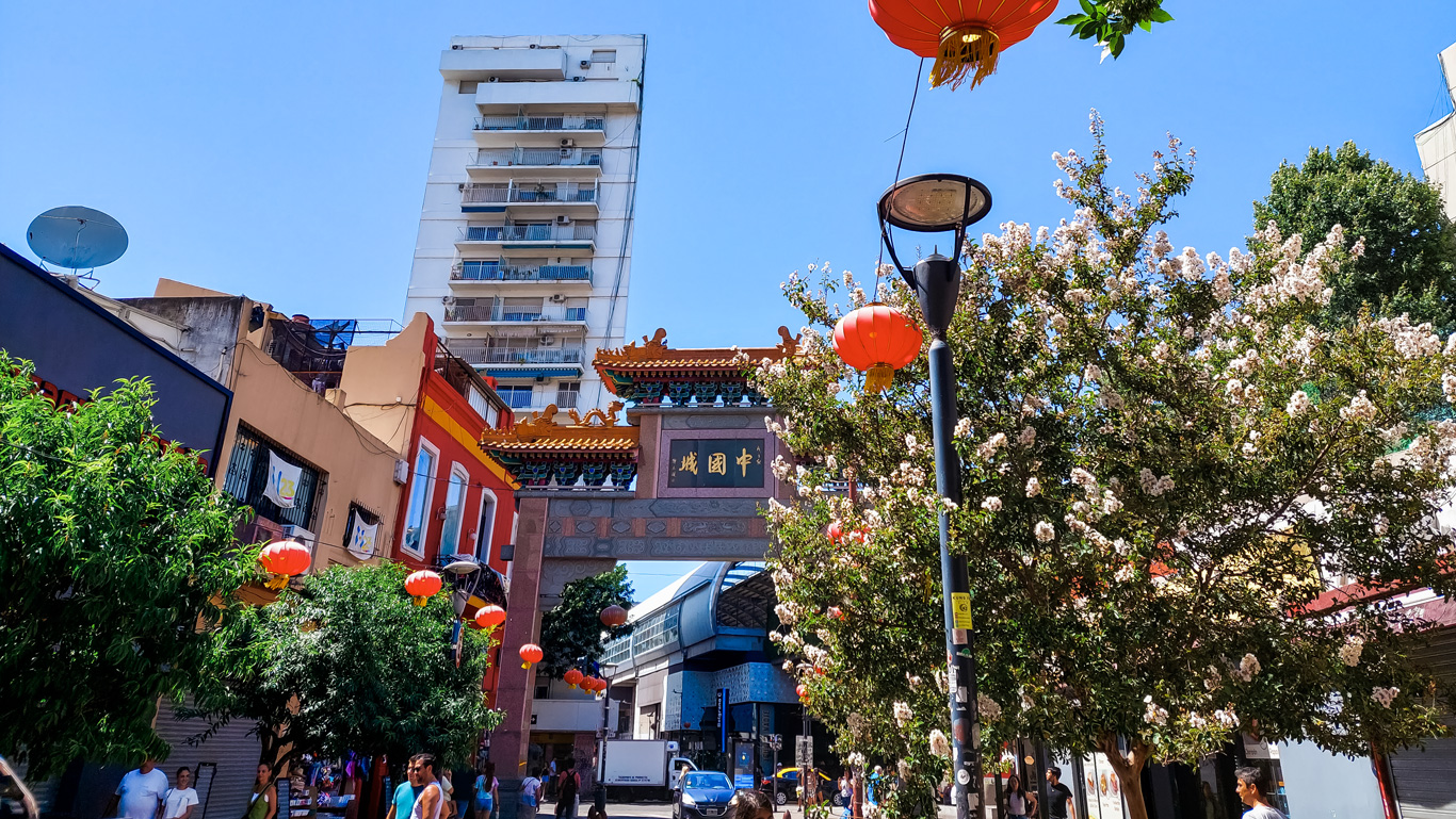 bairro-chino-buenos--aires Entrada do Bairro Chinês de Buenos Aires com portal tradicional, lanternas vermelhas e árvores floridas. Visitantes caminham pelas ruas estreitas em dia de céu azul vibrante.