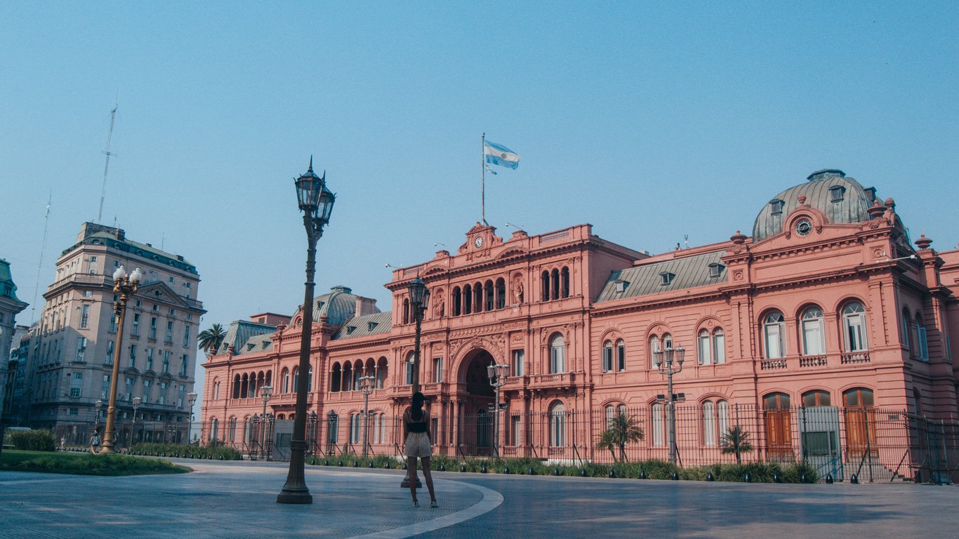 casa-rosada-buenos-aires Fachada da Casa Rosada, sede do governo argentino, em Buenos Aires, com a bandeira da Argentina tremulando no topo. Uma mulher aparece caminhando em frente à praça ampla e vazia.