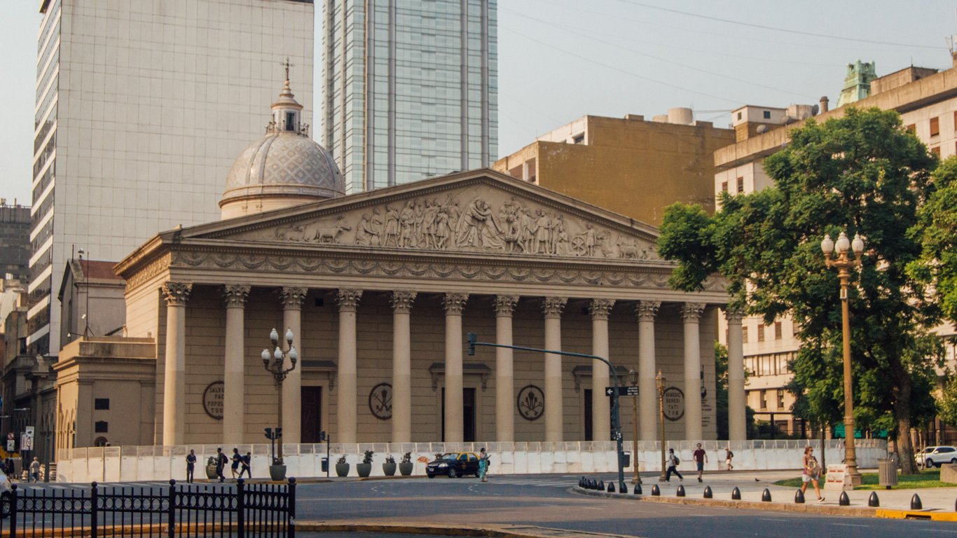 catedral-de-buenos-aires Fachada da Catedral Metropolitana de Buenos Aires com colunas imponentes e esculturas em relevo na parte superior. O prédio histórico contrasta com os edifícios modernos ao fundo.