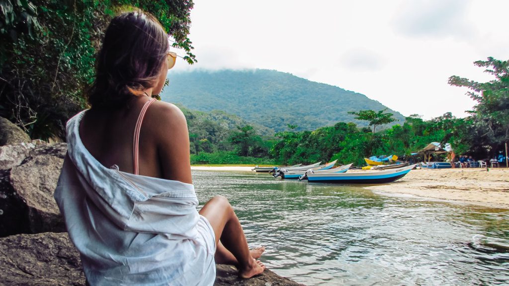 Mulher de costas, sentada sobre uma pedra à beira da água, usando uma camisa branca caída no ombro, observando barcos ancorados em uma praia tranquila cercada por mata atlântica. Ao fundo, montanhas cobertas por névoa completam a paisagem serena — uma cena que remete à tranquilidade de uma viagem costeira, ideal para ilustrar como ir de Ilhabela para Ubatuba explorando paisagens naturais.