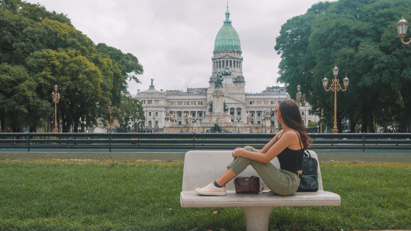 congresso-argentina Mulher sentada em banco de praça observa o prédio do Congresso Nacional, com sua cúpula verde e arquitetura imponente. Árvores envolvem o cenário urbano e institucional.