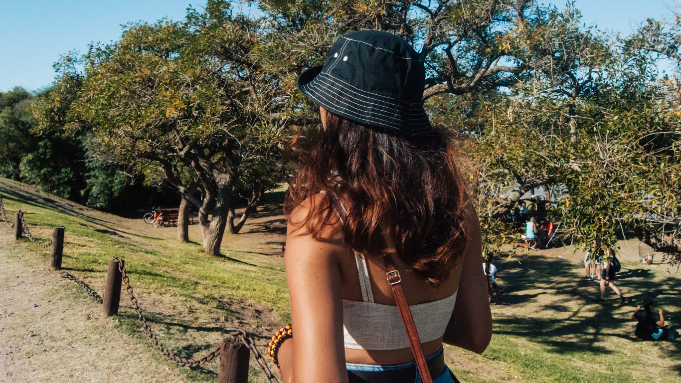 costanera-sur-buenos-aires Uma mulher de chapéu escuro caminha pela Reserva Ecológica Costanera Sur, um parque arborizado em um dia ensolarado, cercada por pessoas relaxando na grama.