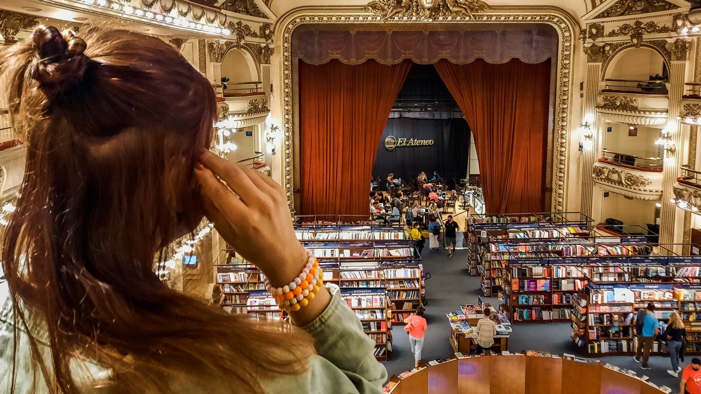 el-ateneo-buenos-aires Mulher observa do mezanino a famosa livraria El Ateneo, instalada em um antigo teatro com cortinas vermelhas no palco e estantes de livros espalhadas pela antiga plateia. O espaço mantém detalhes originais da arquitetura, como ornamentos dourados e varandas internas.