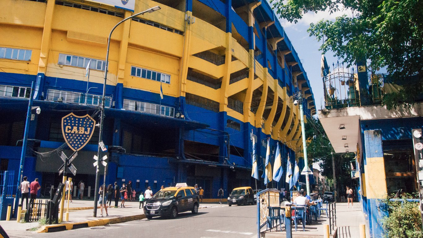 estadio-boca-juniors Fachada do estádio La Bombonera, do Boca Juniors, com torcedores e bandeiras nas cores azul e amarelo.