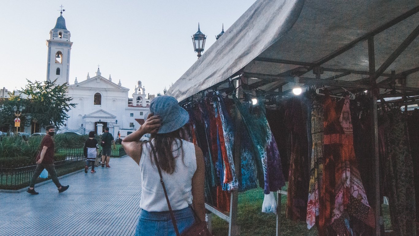 feira-da-recoleta Mulher caminha próxima a barraca de roupas coloridas em feira de artesanato na Recoleta. Ao fundo, a fachada branca da Basílica do Pilar contrasta com o entardecer suave.