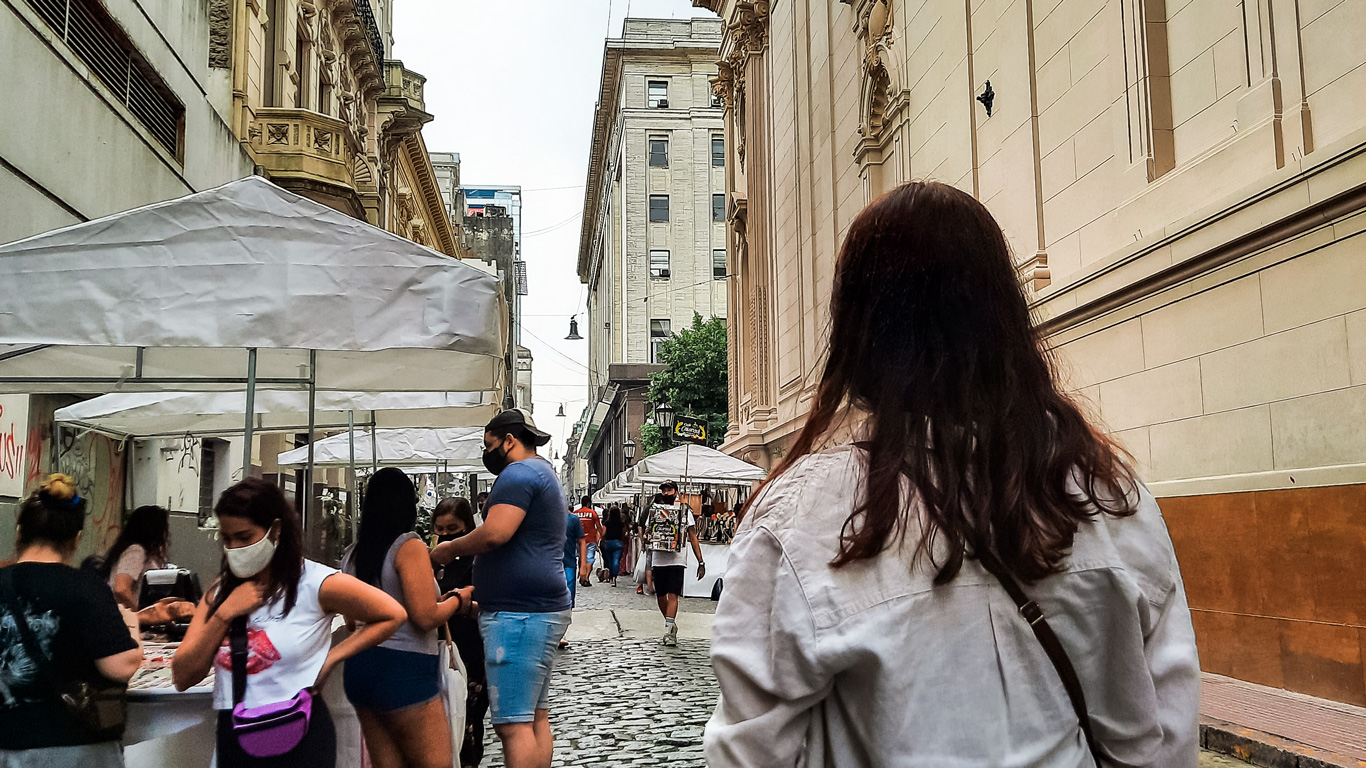 feira-de-san-telmo Mulher caminhando na Rua Defensa em San Telmo, durante a feira de San Telmo, com barracas brancas em ambos os lados, com edifícios históricos ao fundo.