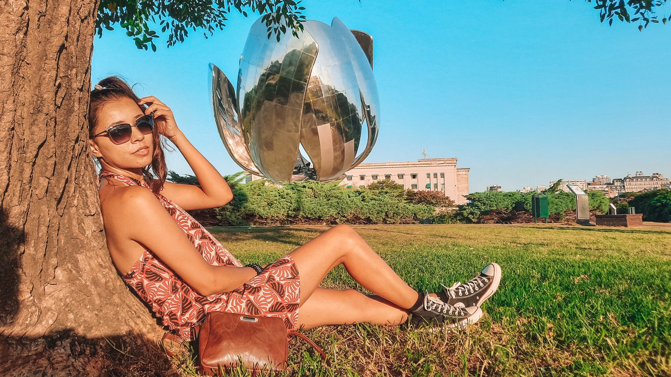 floralis-generica-recoleta Mulher relaxa encostada em uma árvore no parque da Floralis Genérica, escultura metálica em forma de flor localizada em Buenos Aires. O dia ensolarado destaca os tons vibrantes da paisagem.