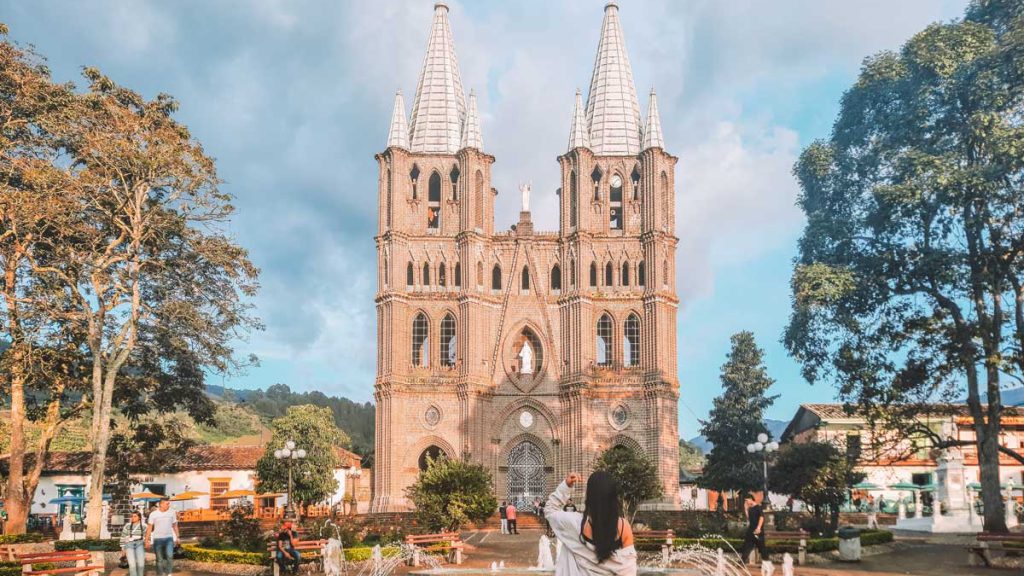 Mulher de costas posa em frente à fonte central da praça principal de Jardín Antioquia, com a imponente Basílica da Imaculada Conceição ao fundo, emoldurada por árvores e céu parcialmente nublado. A arquitetura neogótica da igreja e o clima tranquilo da cidade serrana criam um cenário pitoresco e encantador.