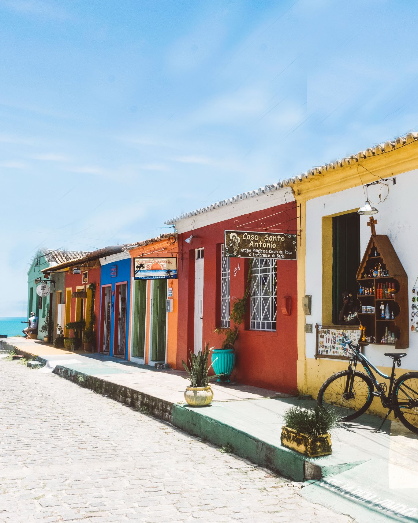 melhor-lugar-para-ficar-em-arraial-dajuda Imagem de uma rua charmosa e tranquila no melhor lugar onde ficar em Arraial d’Ajuda, o centro, com casas coloniais pintadas em cores vibrantes como vermelho, amarelo e azul. Lojas artesanais, bicicletas e vasos com plantas compõem o cenário, com o mar azul ao fundo.