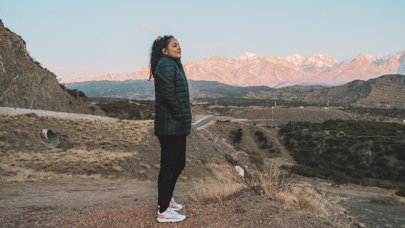 mendoza-em-agosto-4 Mulher vestindo casaco de frio preto e tênis esportivos brancos com rosa posa de perfil em um mirante de terra, com montanhas ao fundo iluminadas pelo sol do fim de tarde. A paisagem árida ao redor, com vegetação rasteira e estrada ao longe, compõe o cenário típico de Mendoza, na Argentina.
