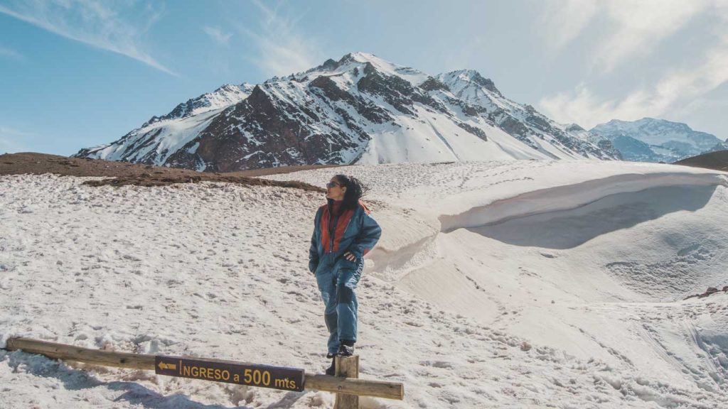 Mulher usando macacão de neve azul com detalhes vermelhos posa em pé sobre uma cerca de madeira, com uma placa que diz “INGRESO a 500 mts.” ao fundo. Ela está cercada por um cenário montanhoso coberto de neve, com picos elevados e céu claro, provavelmente em uma região andina da Argentina no inverno.