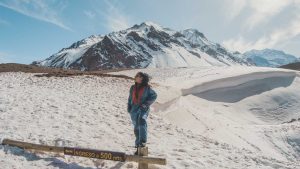 Mulher usando macacão de neve azul com detalhes vermelhos posa em pé sobre uma cerca de madeira, com uma placa que diz “INGRESO a 500 mts.” ao fundo. Ela está cercada por um cenário montanhoso coberto de neve, com picos elevados e céu claro, provavelmente em uma região andina da Argentina no inverno.