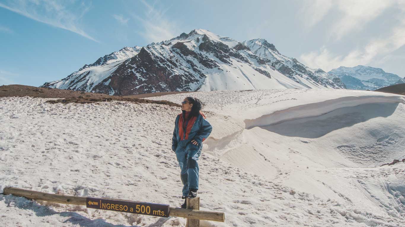 Mulher usando macacão de neve azul com detalhes vermelhos posa em pé sobre uma cerca de madeira, com uma placa que diz “INGRESO a 500 mts.” ao fundo. Ela está cercada por um cenário montanhoso coberto de neve, com picos elevados e céu claro, provavelmente em uma região andina da Argentina no inverno.