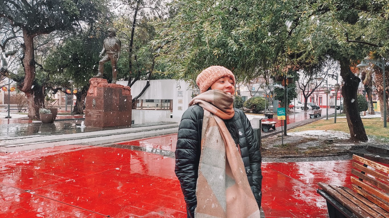 mendoza-em-agosto-neve Mulher sorridente com touca e cachecol em uma praça de Mendoza enquanto flocos de neve caem; ao fundo, uma estátua e árvores cobertas parcialmente de neve.