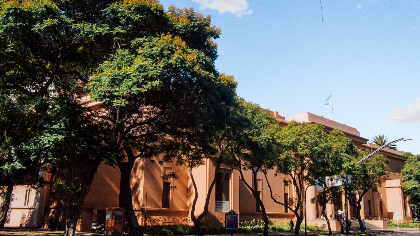 museu-nacional-de-belas-artes Fachada do Museu Nacional de Belas Artes, cercada por árvores e vegetação densa. O prédio em tom terracota exibe bandeira da Argentina e se destaca sob céu azul com poucas nuvens.