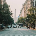 Avenida que leva ao Obelisco de Buenos Aires, um monumento icônico da Argentina e uma das melhores opções de o que fazer em Buenos Aires. A rua é ladeada por prédios altos e árvores, e várias pessoas estão atravessando a faixa de pedestres, com tráfego leve visível. O Obelisco se destaca ao fundo.
