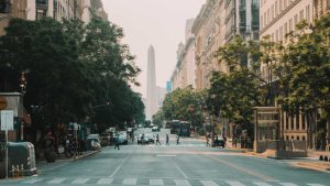 Avenida que leva ao Obelisco de Buenos Aires, um monumento icônico da Argentina e uma das melhores opções de o que fazer em Buenos Aires. A rua é ladeada por prédios altos e árvores, e várias pessoas estão atravessando a faixa de pedestres, com tráfego leve visível. O Obelisco se destaca ao fundo.