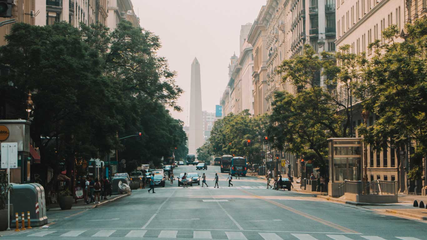 Avenida que leva ao Obelisco de Buenos Aires, um monumento icônico da Argentina e uma das melhores opções de o que fazer em Buenos Aires. A rua é ladeada por prédios altos e árvores, e várias pessoas estão atravessando a faixa de pedestres, com tráfego leve visível. O Obelisco se destaca ao fundo.