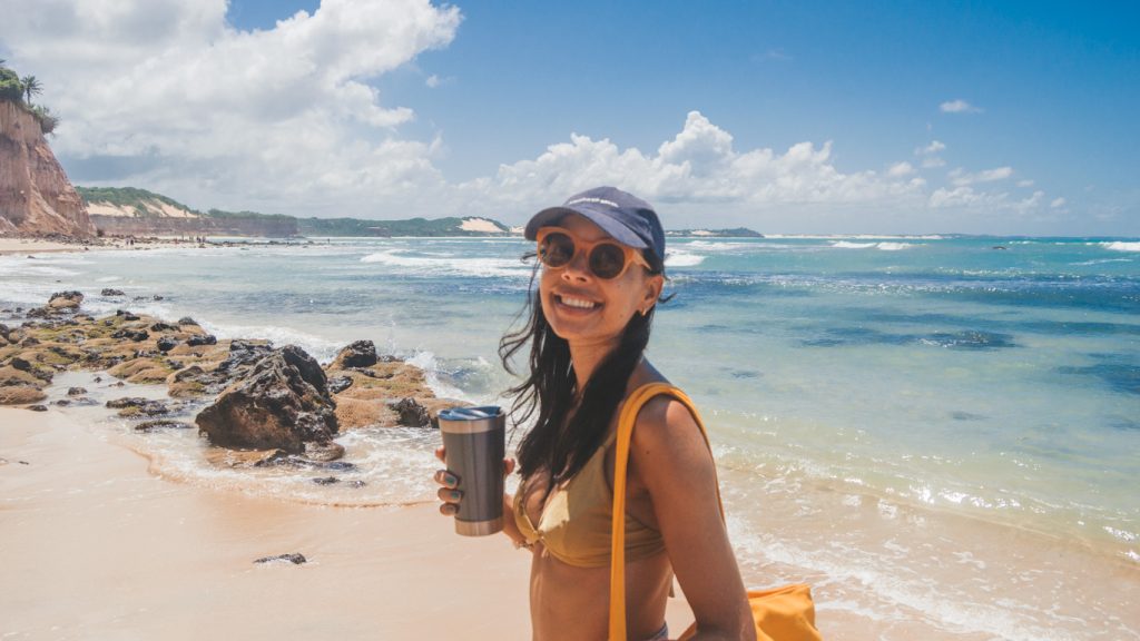 Mulher sorridente de biquíni amarelo, óculos de sol e boné, segurando uma garrafa térmica na Praia de Pipa com mar azul, céu ensolarado e falésias ao fundo.