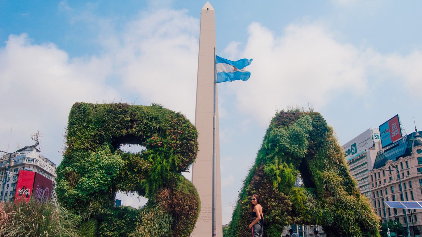 obelisco-de-buenos-aires-1 Mulher posa entre as esculturas vegetais em forma de "BA", em frente ao Obelisco de Buenos Aires. Ao fundo, a bandeira da Argentina tremula sob um céu parcialmente nublado.