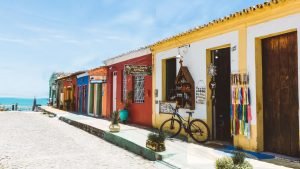 Imagem de uma rua charmosa e tranquila no melhor lugar onde ficar em Arraial d’Ajuda, o centro, com casas coloniais pintadas em cores vibrantes como vermelho, amarelo e azul. Lojas artesanais, bicicletas e vasos com plantas compõem o cenário, com o mar azul ao fundo.