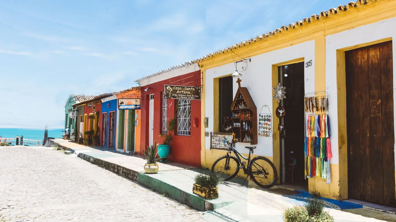 onde-ficar--em-arraial-dajuda Imagem de uma rua charmosa e tranquila no melhor lugar onde ficar em Arraial d’Ajuda, o centro, com casas coloniais pintadas em cores vibrantes como vermelho, amarelo e azul. Lojas artesanais, bicicletas e vasos com plantas compõem o cenário, com o mar azul ao fundo.