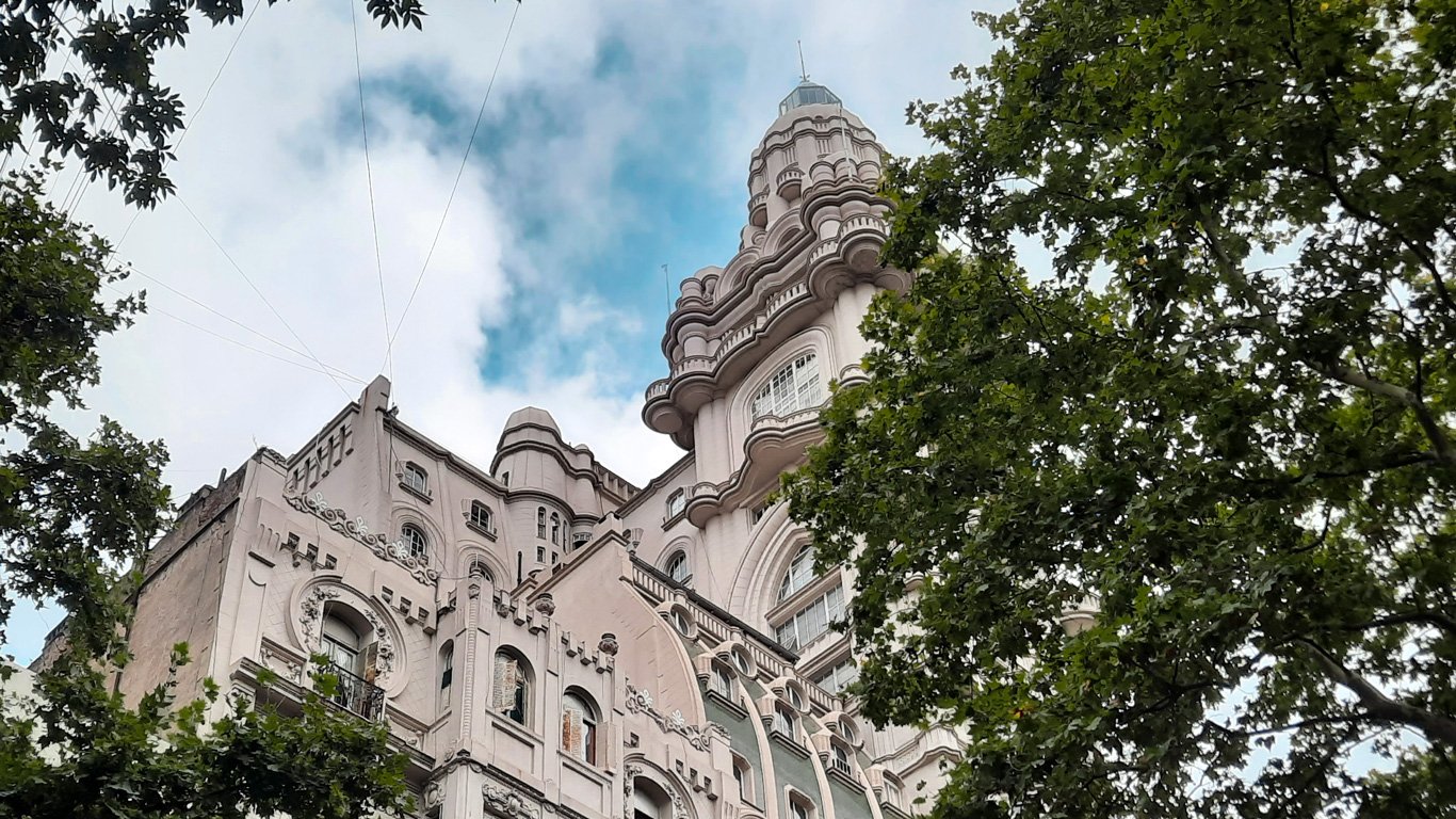 palacio-barolo Vista de baixo do Palácio Barolo, com sua arquitetura detalhada e torres inspiradas na Divina Comédia. Árvores emolduram a construção imponente sob um céu parcialmente nublado.