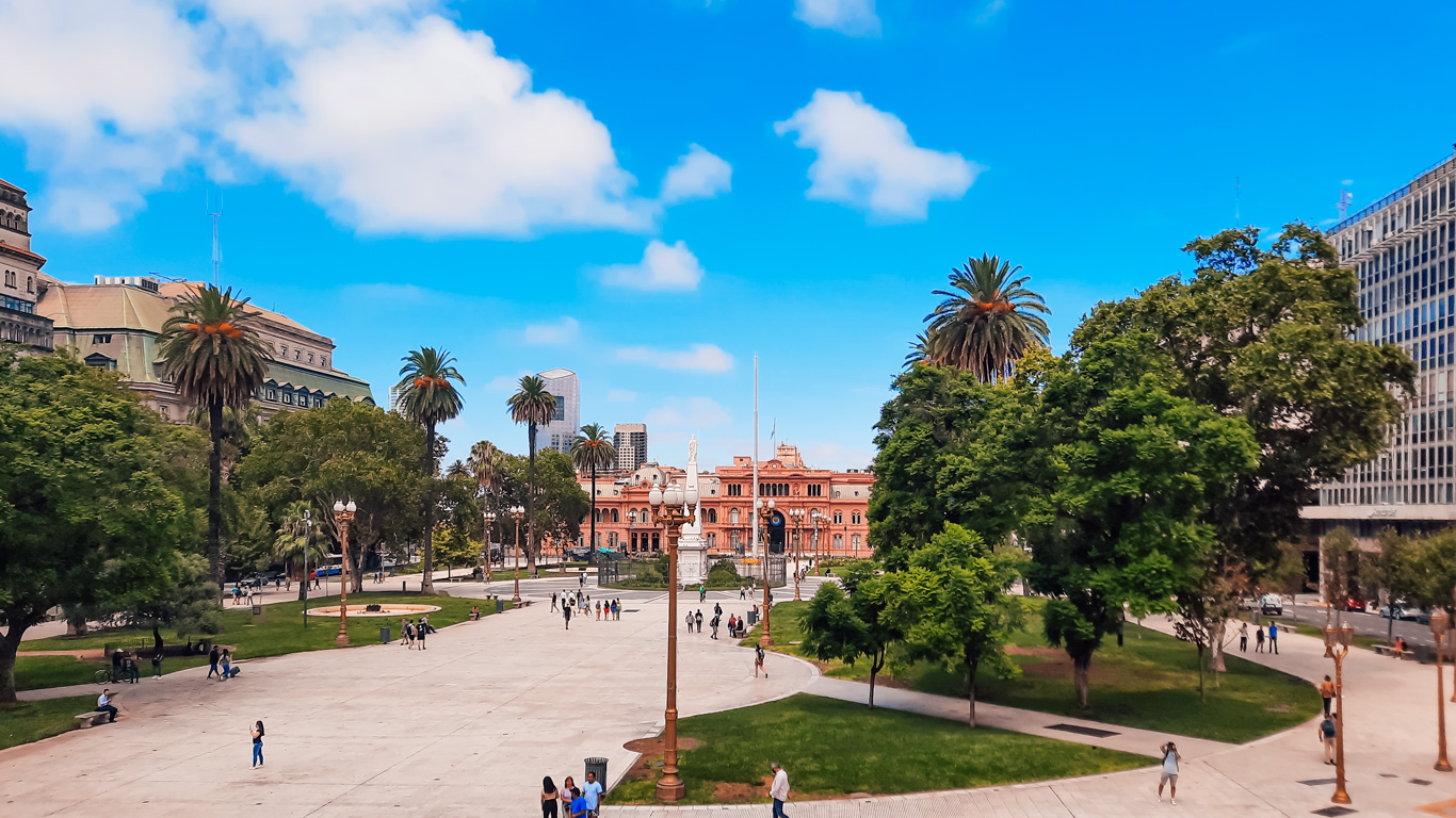 plaza-de-mayo-2 ista ampla da Plaza de Mayo com a Casa Rosada ao fundo, cercada por palmeiras e prédios históricos. Pessoas caminham pela praça sob um céu azul vibrante.