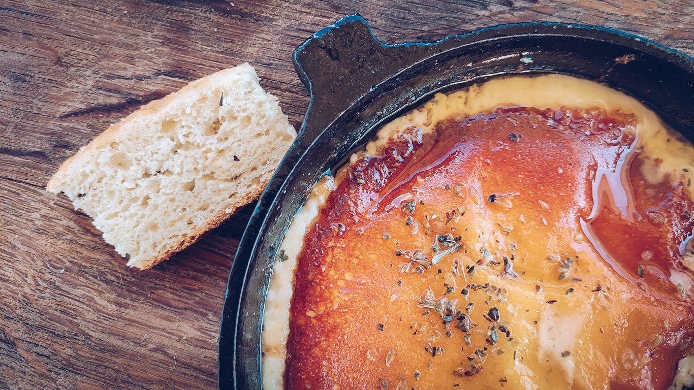 provolleta-culinaria-argentina-1 Close de uma provoleta derretida coberta com orégano em frigideira de ferro, acompanhada de um pedaço de pão rústico sobre mesa de madeira.