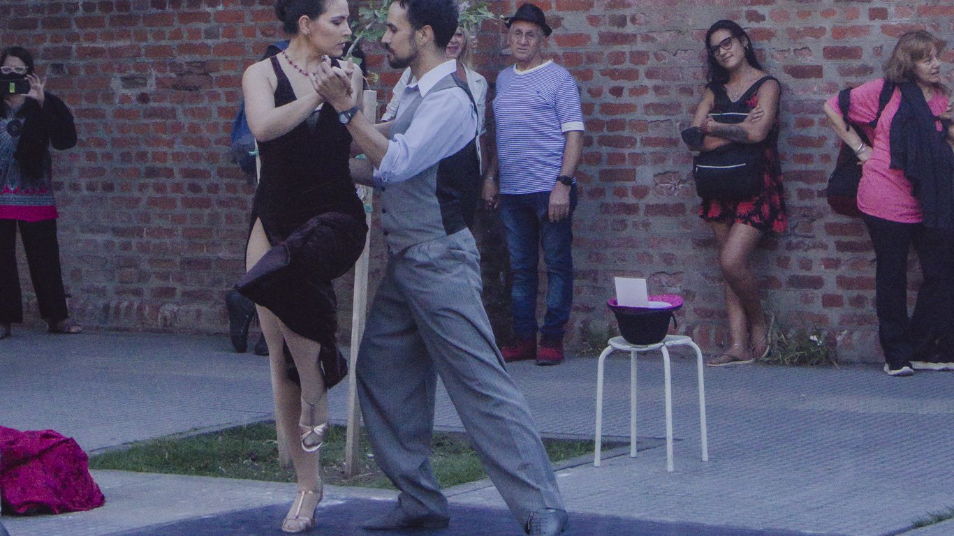 tango-em-buenos-aires Casal se apresenta com look clássico de tango nas ruas de Buenos Aires; em frente ao Cemitério da Recoleta, ela usa vestido preto com fenda e salto, enquanto ele veste camisa e colete cinza. Pessoas observam ao fundo a performance intensa e apaixonada.