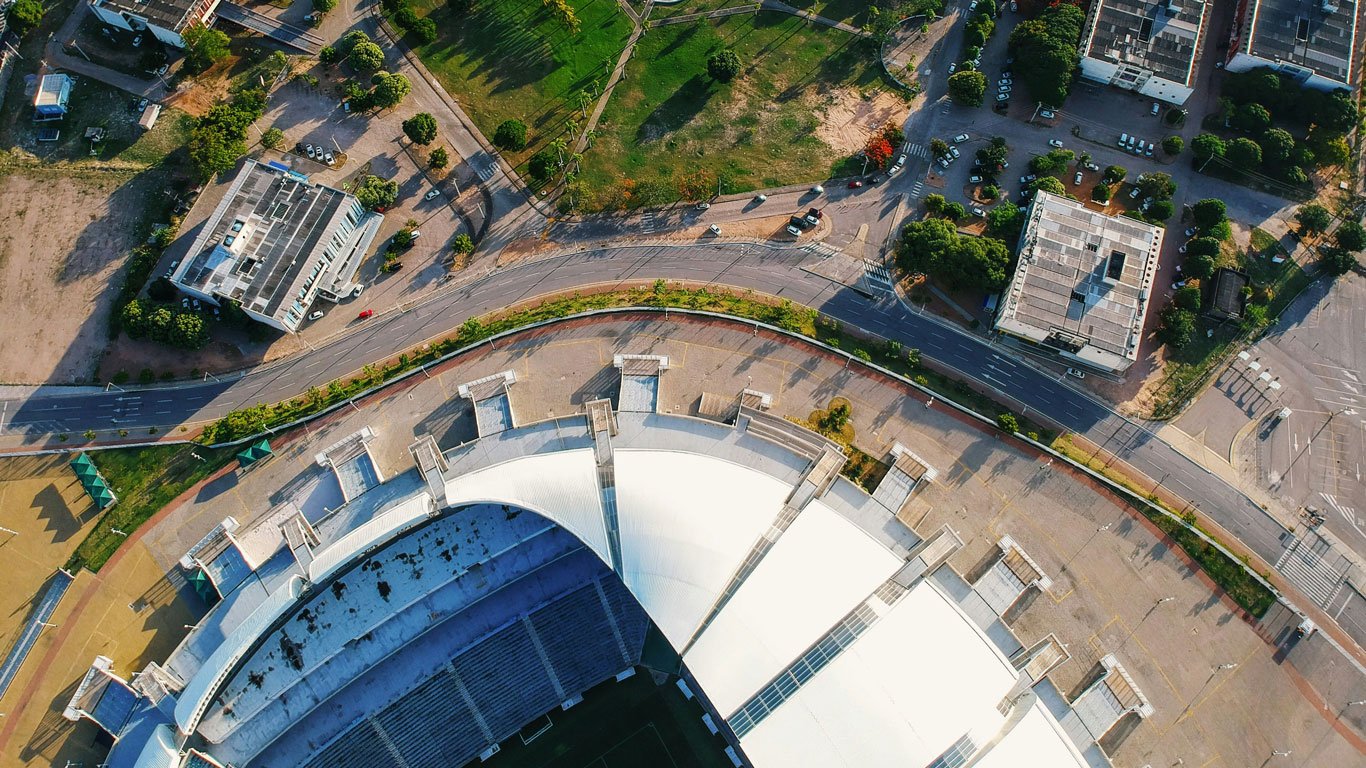 arena-das-dunas-natal Imagem aérea mostrando parte superior do estádio Arena das Dunas, em Natal, RN, com seu telhado branco curvo em destaque e ruas e prédios ao redor, incluindo áreas verdes com caminhos sinuosos.