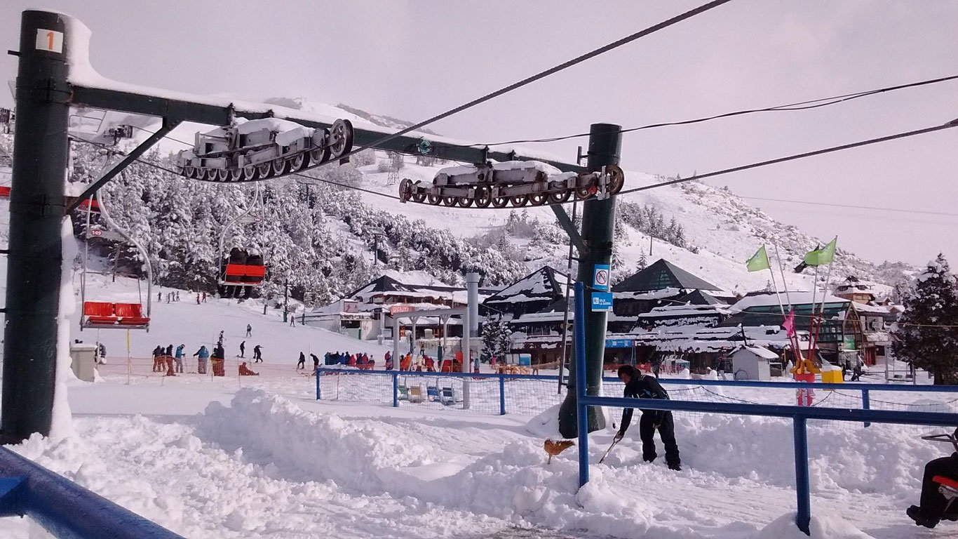 aulas-de-ski-em-bariloche Cena de uma estação de esqui movimentada no Cerro Catedral, em Bariloche, com turistas e instrutores praticando esqui na neve. Há teleféricos em funcionamento e prédios cobertos de neve ao fundo, indicando um típico dia de inverno.