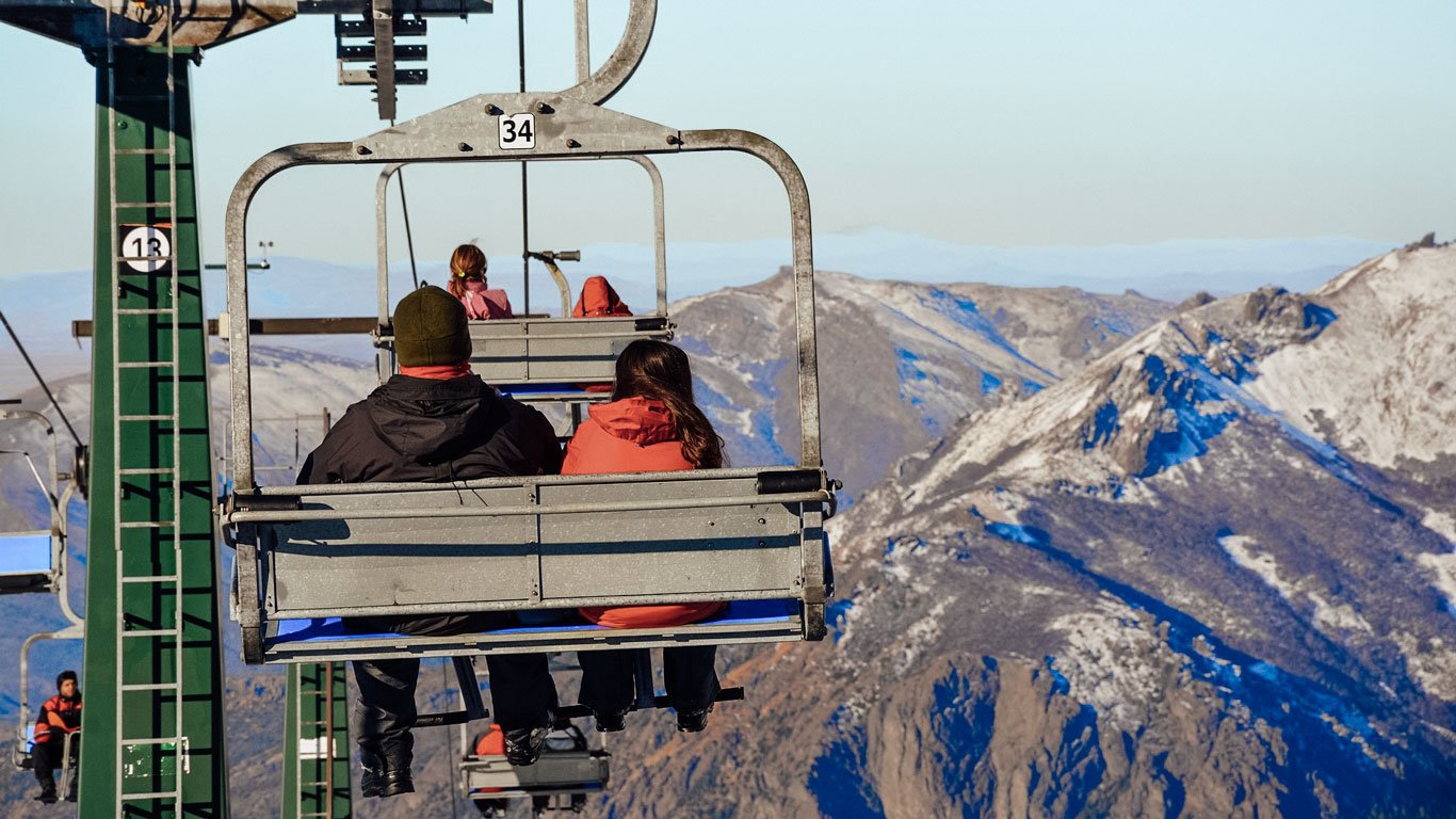 bariloche-no-inverno-roupas Pessoas sentadas em cadeiras de teleférico subindo o Cerro em Bariloche, com montanhas rochosas e parcialmente nevadas ao fundo.