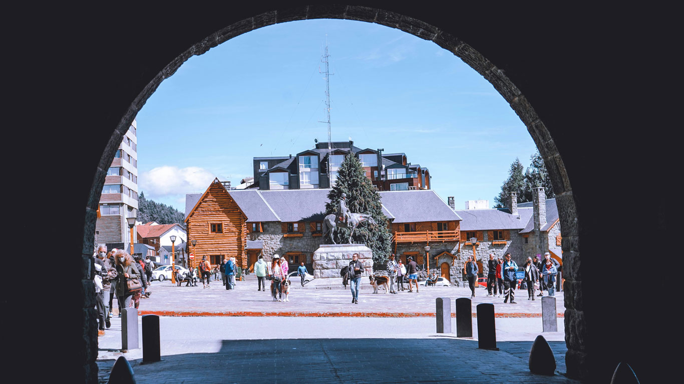 bariloche-no-inverno Fotografia tirada de um arco de pedra que emoldura o famoso Centro Cívico de Bariloche. Pessoas caminham pela praça em frente à estátua de San Martín, cercada por construções de madeira e pedra, sob um céu azul claro.