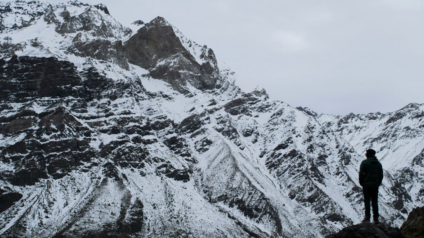 cajon-del-maipo-chile Pessoa em pé sobre uma rocha em Cajón del Maipo, observa montanhas cobertas de neve sob um céu nublado, destacando a imponência da Cordilheira dos Andes.