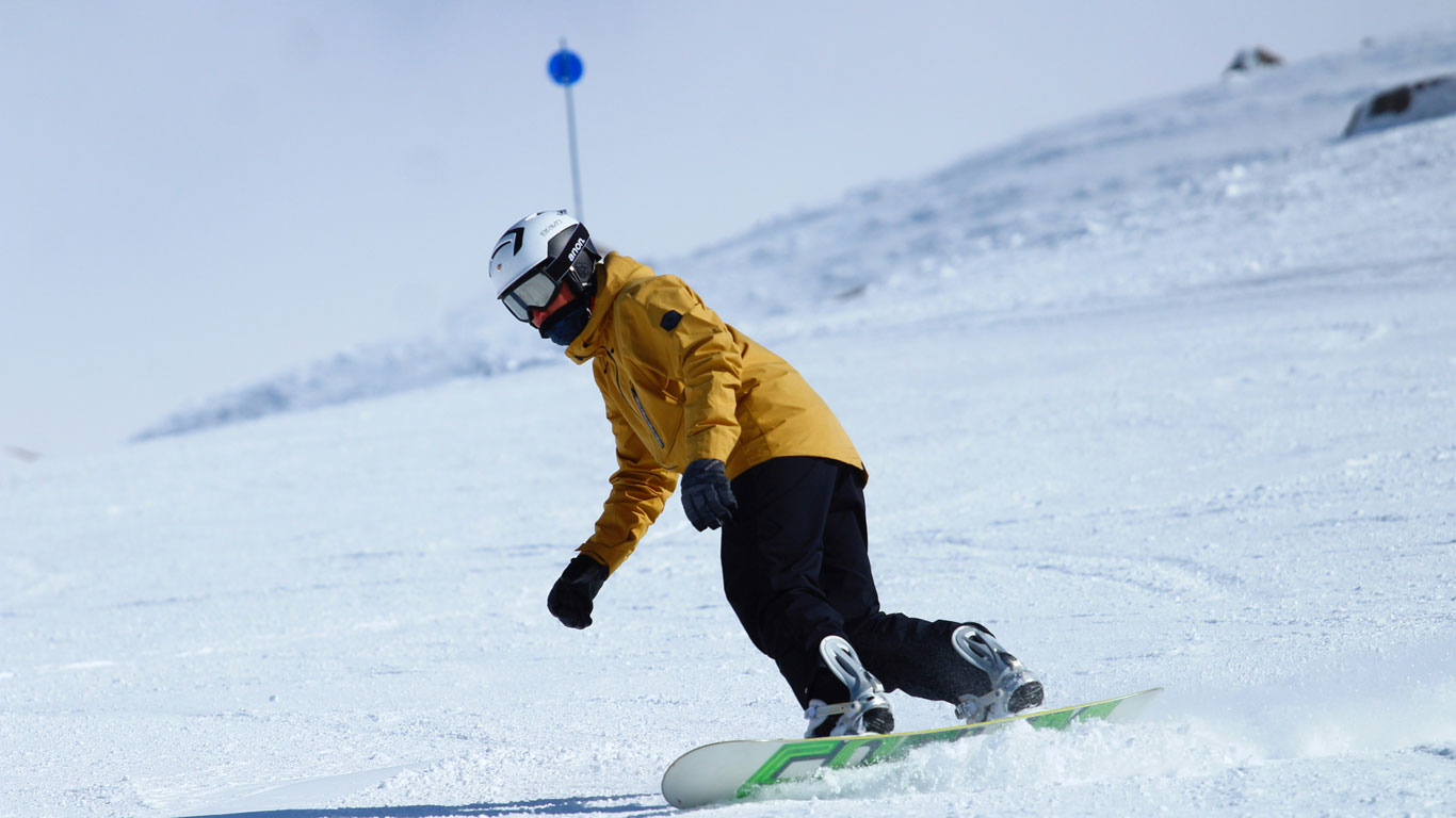 cerro-catedral-bariloche Pessoa praticando snowboard em uma encosta nevada no Cerro Catedral, em Bariloche. Ela veste jaqueta amarela, calça preta, capacete branco e óculos de proteção, deslizando com habilidade sobre a neve sob um céu claro.