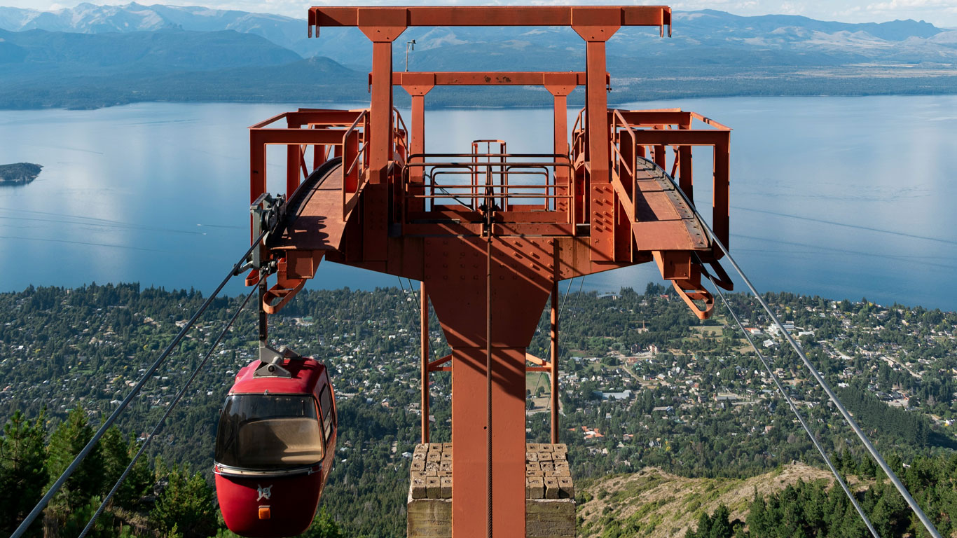 cerro-otto-bariloche Imagem de um bondinho vermelho suspenso no ar no Cerro Otto, com vista panorâmica de Bariloche e do Lago Nahuel Huapi. A estrutura metálica se destaca no topo da montanha, cercada por florestas e casas ao longe.
