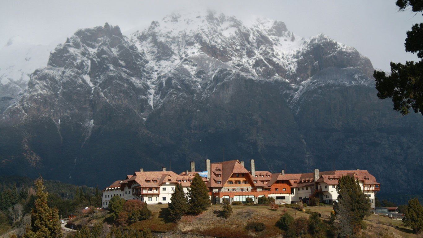 circuito-chico-bariloche Hotel Llao Llao em destaque, com arquitetura charmosa de telhado vermelho e paredes claras, posicionado diante de uma imensa montanha nevada.