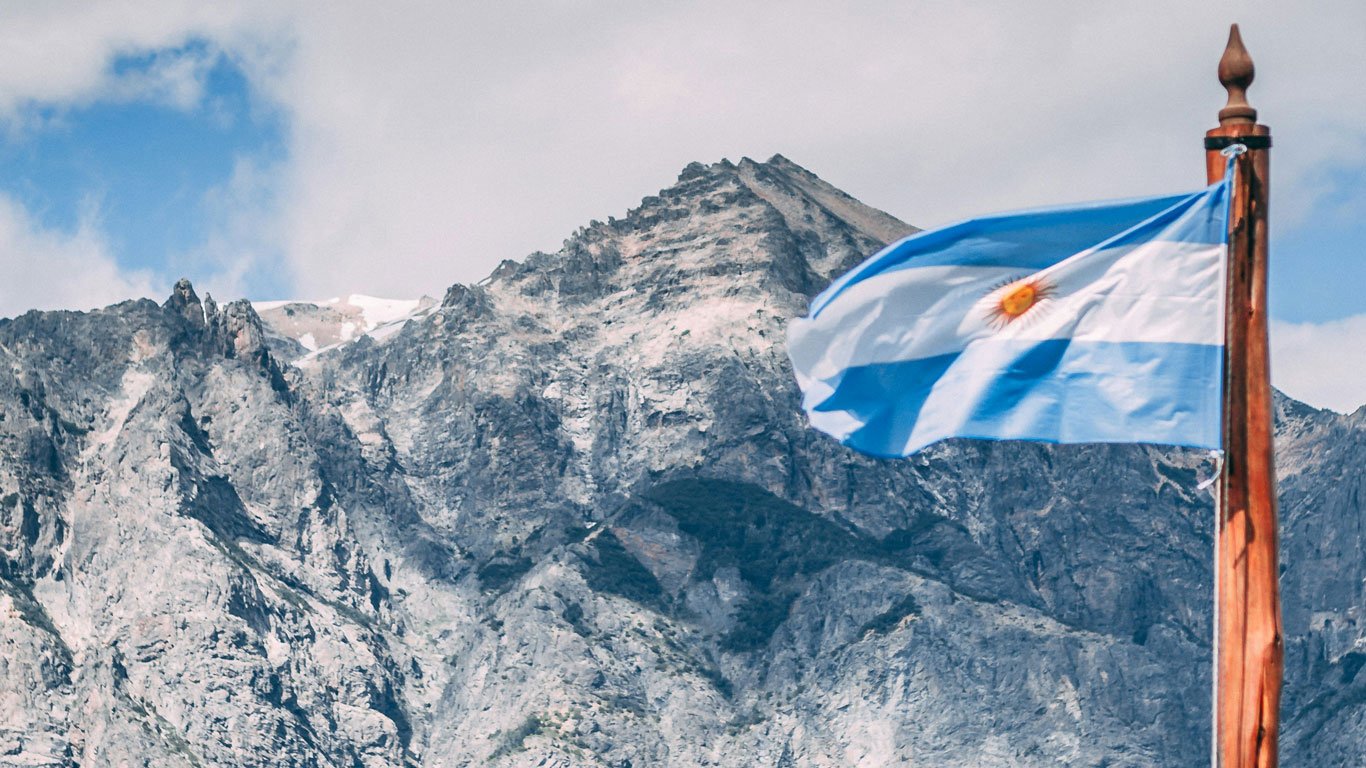 clima-em-bariloche-no-inverno Bandeira da Argentina tremulando ao vento em frente a uma imponente cadeia de montanhas rochosas. O céu está parcialmente nublado, e o topo da montanha exibe traços de neve, sugerindo clima frio típico de Bariloche no inverno.