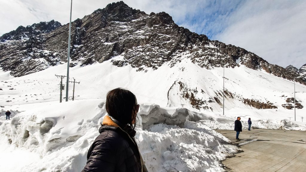 Mulher de casaco e óculos escuros admira uma montanha com neve em Santiago, Na Cordilheira dos Andes, enquanto outras pessoas caminham sobre uma estrada parcialmente limpa de neve.