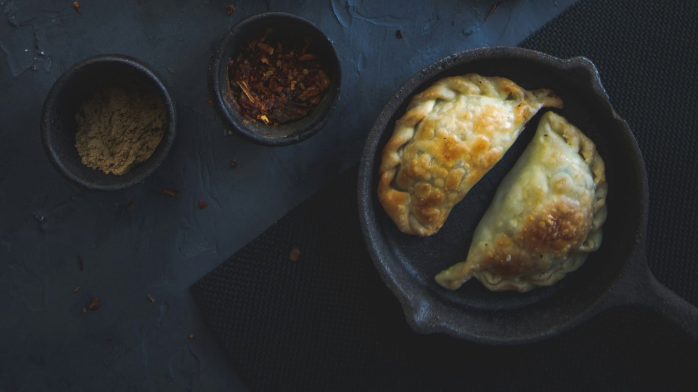 culinaria-ushuaia Empanadas argentinas douradas servidas em frigideira de ferro ao lado de potes com especiarias secas, sobre superfície escura.