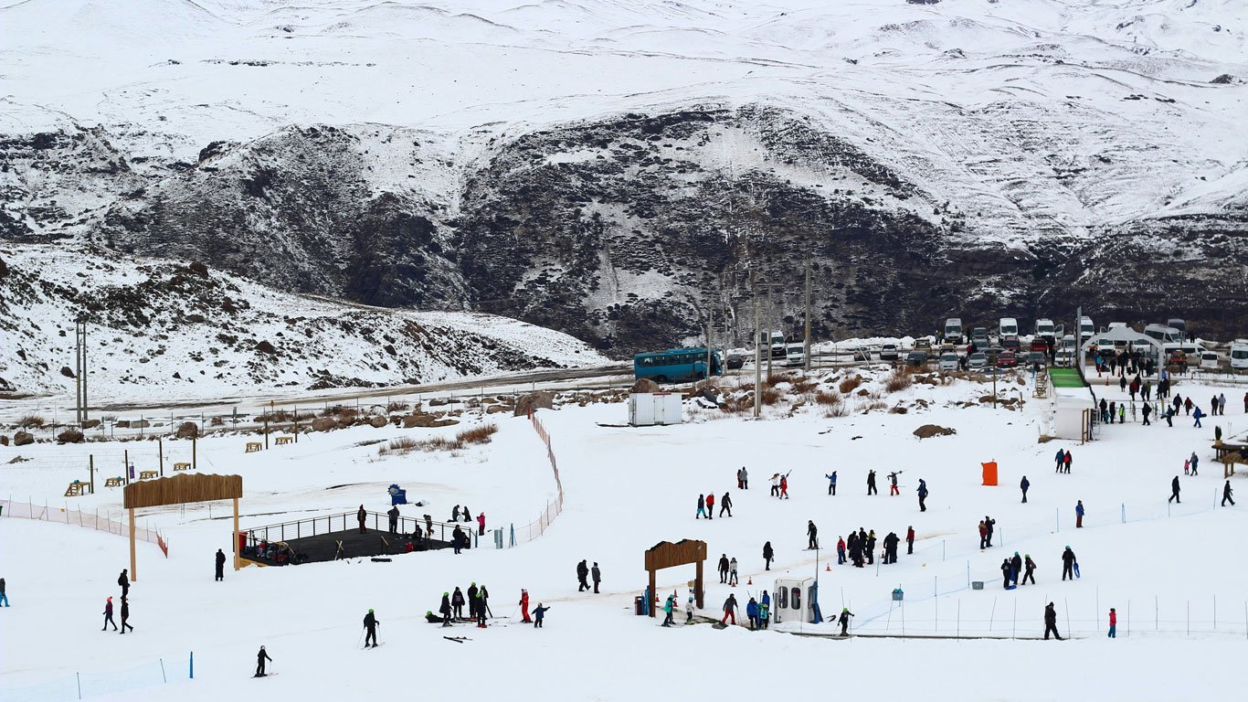farellones-chile Diversas pessoas se divertem esquiando e caminhando sobre uma grande área coberta de neve em Farellones, com ônibus e carros estacionados ao fundo.