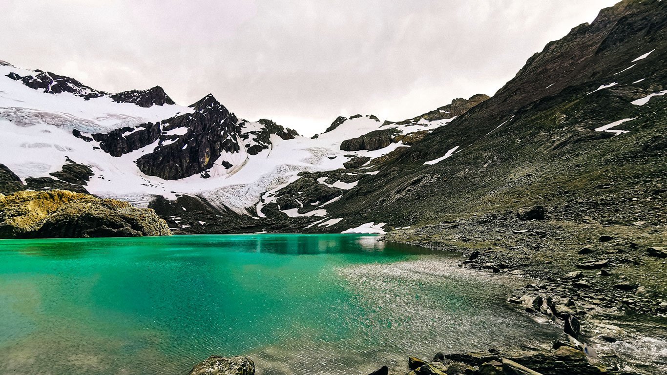 glaciar-Vinciguerra-Ushuaia Lago de águas verde-esmeralda refletindo a luz sob céu nublado, rodeado por encostas rochosas e o Glaciar Vinciguerra parcialmente coberto de neve nos arredores de Ushuaia.