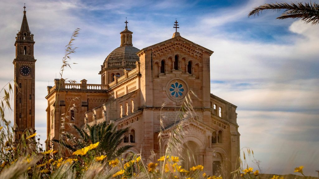 gozo-malta-1 A Basílica de Ta' Pinu, em Gozo, ergue-se majestosamente em meio às flores silvestres, com sua arquitetura ornamentada e torres sineiras gêmeas se estendendo em direção a um céu azul suave, emanando uma sensação de paz e grandiosidade espiritual.