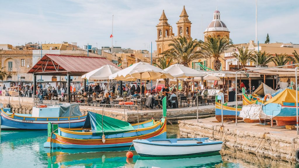 marsalokk Cena movimentada à beira-mar em Marsaxlokk, em Malta, com os tradicionais barcos de pesca malteses coloridos, conhecidos como'luzzus', flutuando nas águas azuis cristalinas. O fundo apresenta cafés ao ar livre movimentados com pessoas sob guarda-sóis brancos e arquitetura histórica, incluindo as icônicas torres da igreja.