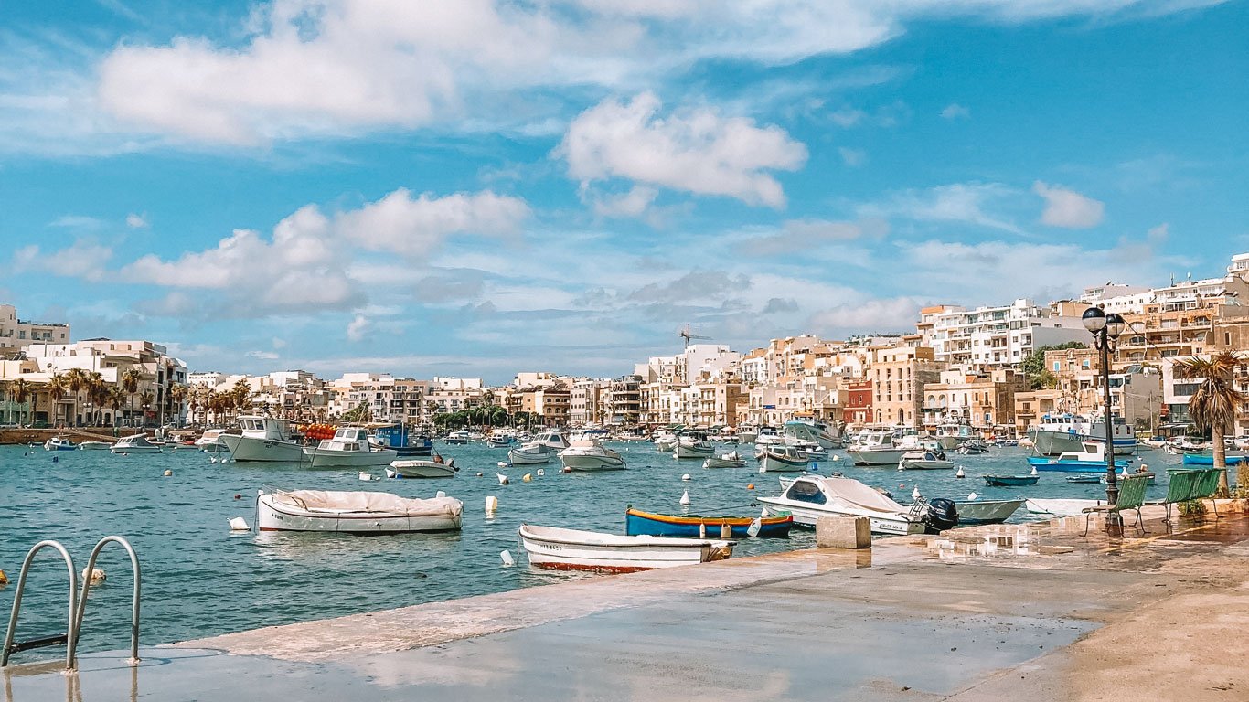 marsaskala-malta O mar azul de Marsaskala, em um dia de sol, com os barcos coloridos ancorados, ao fundo as casas e pequenos edifícios de tons claros compõem a paisagem.