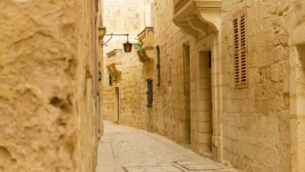 mdina-malta Uma rua estreita e tranquila na antiga cidade murada de Mdina, Malta, caracterizada pelas suas paredes de calcário, portas em arco e varandas tradicionais maltesas com persianas de madeira fechadas. O encanto intemporal da cidade é realçado pelos tons quentes da pedra e pelo verde ocasional dos vasos de plantas.