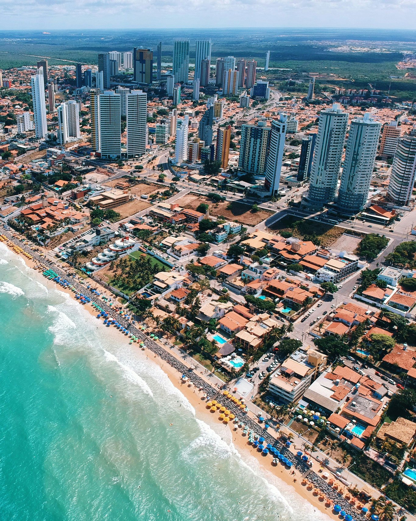 melhor-lugar-para-ficar-em-natal Vista aérea de Ponta Negra, o melhor bairro onde ficar em Natal. Com prédios modernos contrastando com as casas de praia, praias movimentadas e guarda-sóis coloridos alinhados na orla. O mar azul-turquesa contrasta com a urbanização intensa e o verde da vegetação ao fundo.